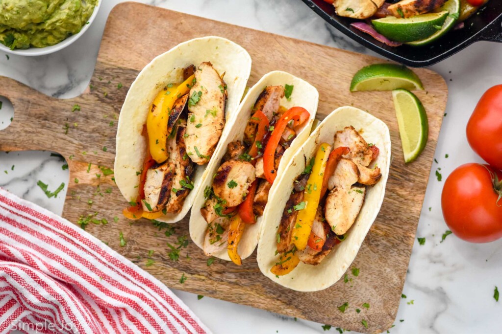 overhead view of chicken fajitas in tortillas on wooden serving board with lime wedges sitting beside.