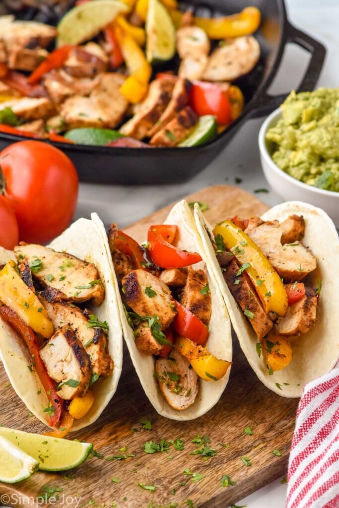 Chicken Fajitas in tortillas with bowl of guacamole and skillet of chicken fajitas sitting in background