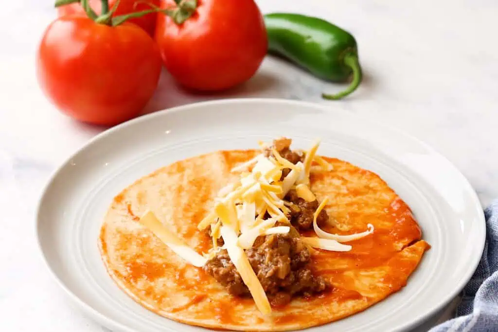 adding ground beef and cheese to a dipped corn tortilla to make enchiladas