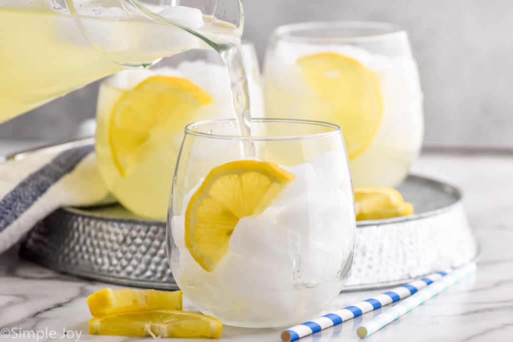 Side view of pitcher of lemonade being poured into glass of ice and lemon wedge for Vodka Lemonade recipe. More glasses of Vodka Lemonade in the background. Straws and lemon wedges next to glass.