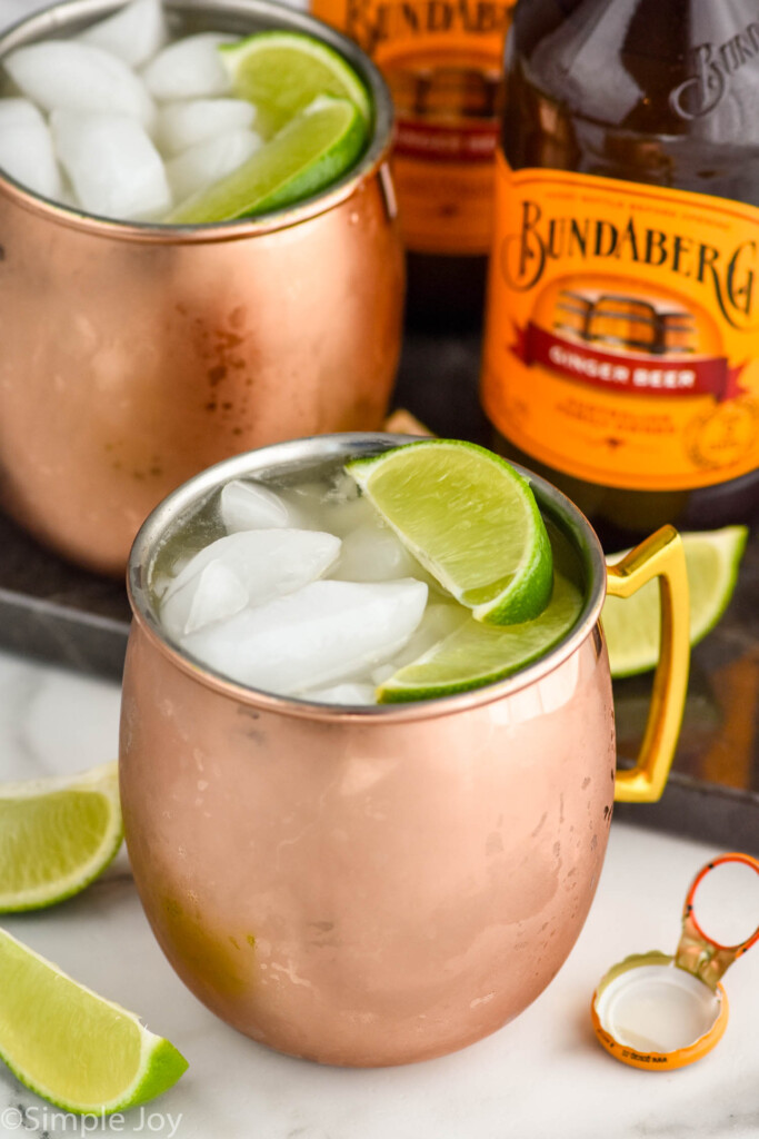 Side view of copper mugs of tequila mules recipe and lime wedges for garnish. Two bottles of ginger beer behind copper mugs. Extra lime wedges and a ginger beer bottle cap on counter.