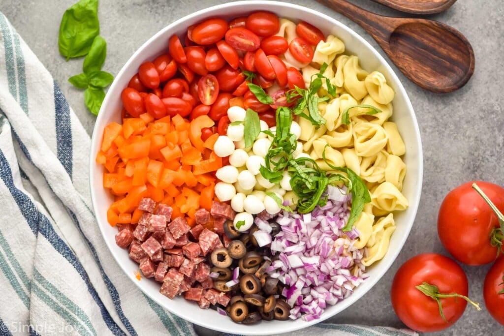 Overhead photo of a bowl of ingredients for Tortellini Pasta Salad recipe. Serving spoons and tomatoes beside bowl.