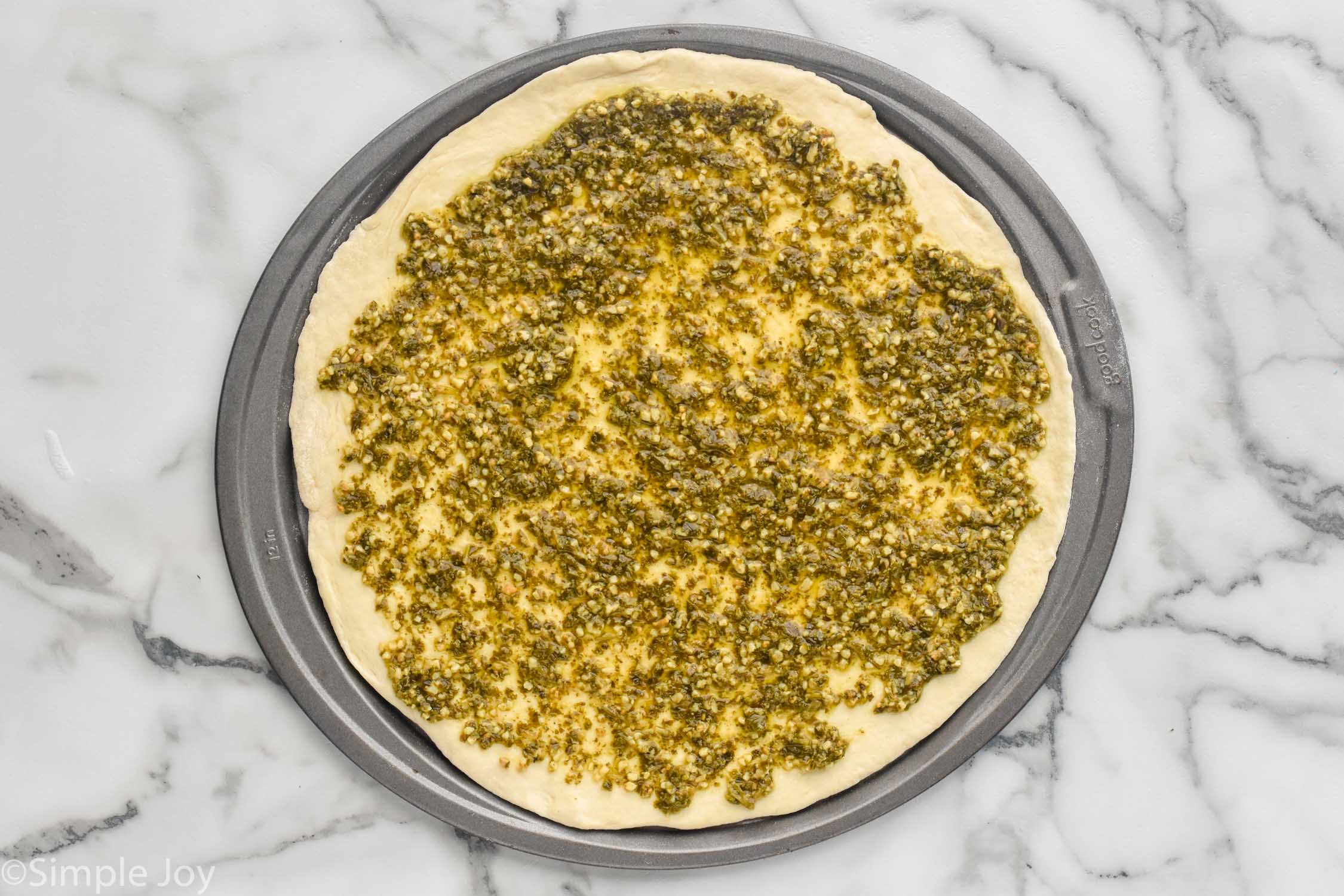 Overhead photo of pizza crust and pesto on baking sheet for Pesto Pizza recipe.