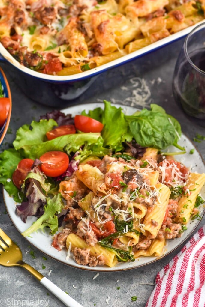 Photo of a plate of Baked Rigatoni and side salad with a fork. Baking dish of Baked Rigatoni and a glass of wine beside plate.