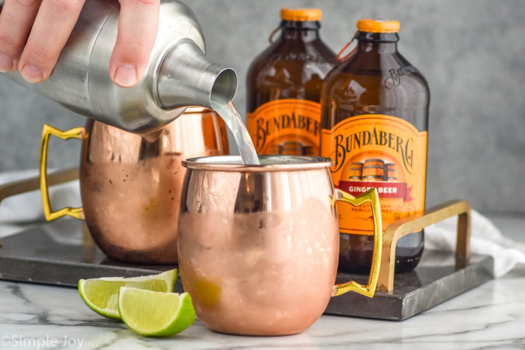 Side view of person's hand pouring a cocktail shaker of ingredients into a copper mug for tequila mules recipe. Lime wedges on counter for garnish, Another copper mug and two bottles of ginger beer behind front cup.
