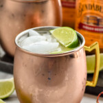Side view of copper mugs of tequila mules garnished with lime wedges and ice. Extra lime wedges on counter beside copper mugs. Bottles of ginger beer beside copper mugs.