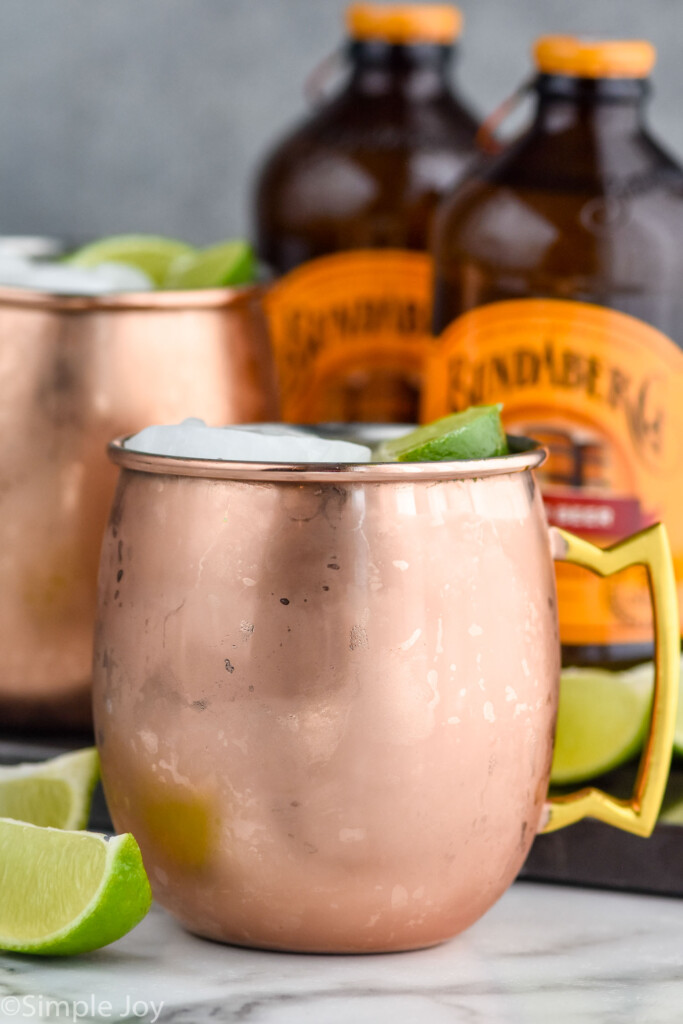 Side view of copper mugs of tequila mules recipe and lime wedges for garnish. Two bottles of ginger beer behind copper mugs.