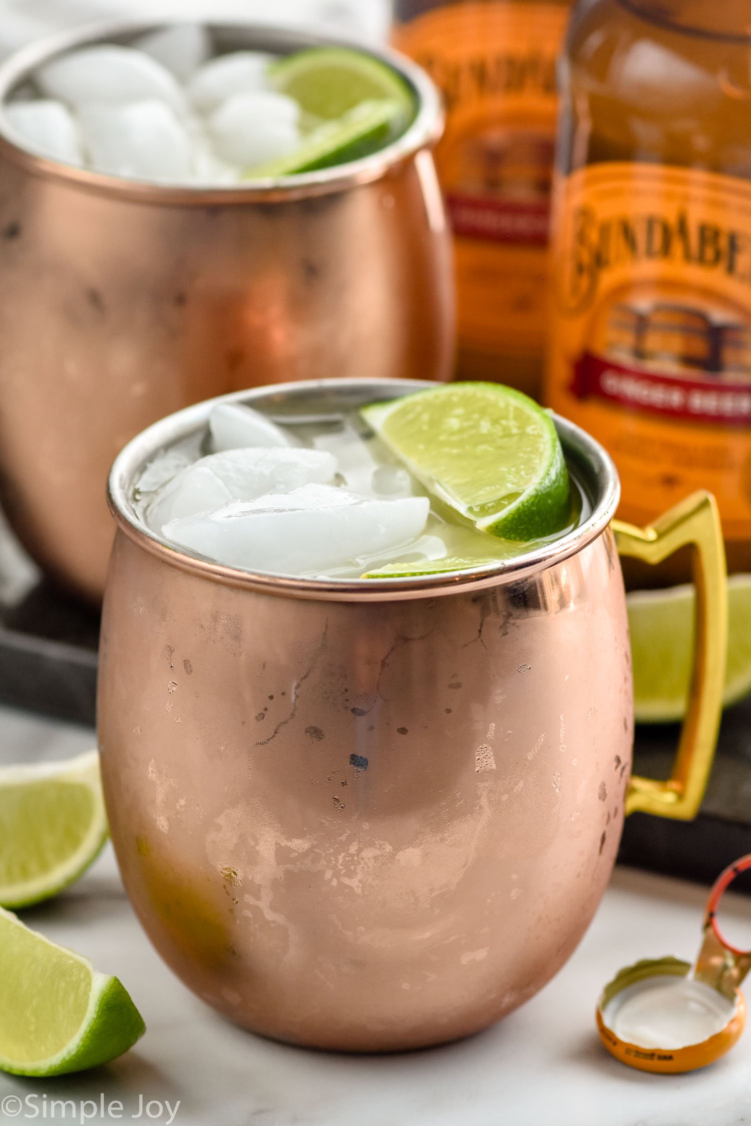 Side view of copper mugs of tequila mules garnished with lime wedges and ice. Extra lime wedges on counter beside copper mugs. Bottles of ginger beer beside copper mugs.