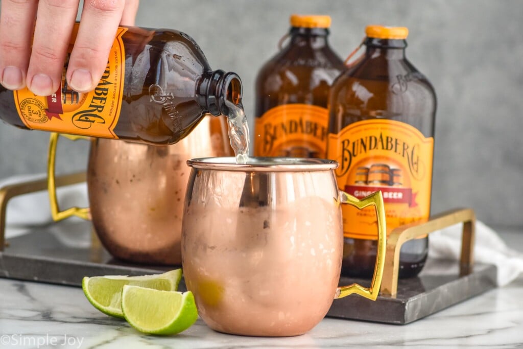 Side view of person's hand pouring ginger beer into a copper mug. More ginger beer and copper mug in the background. Lime wedges beside copper mug for garnish.