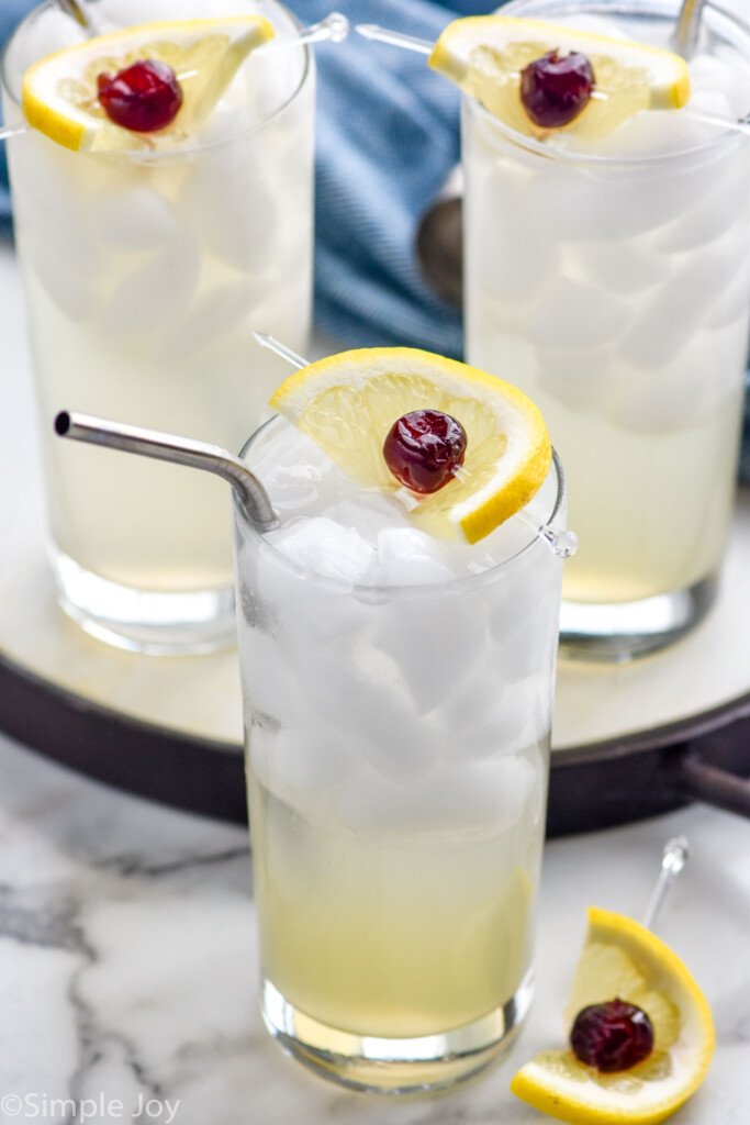 Three glasses of Tom Collins cocktails with ice, a straw, and garnished with a lemon wedge and cherry