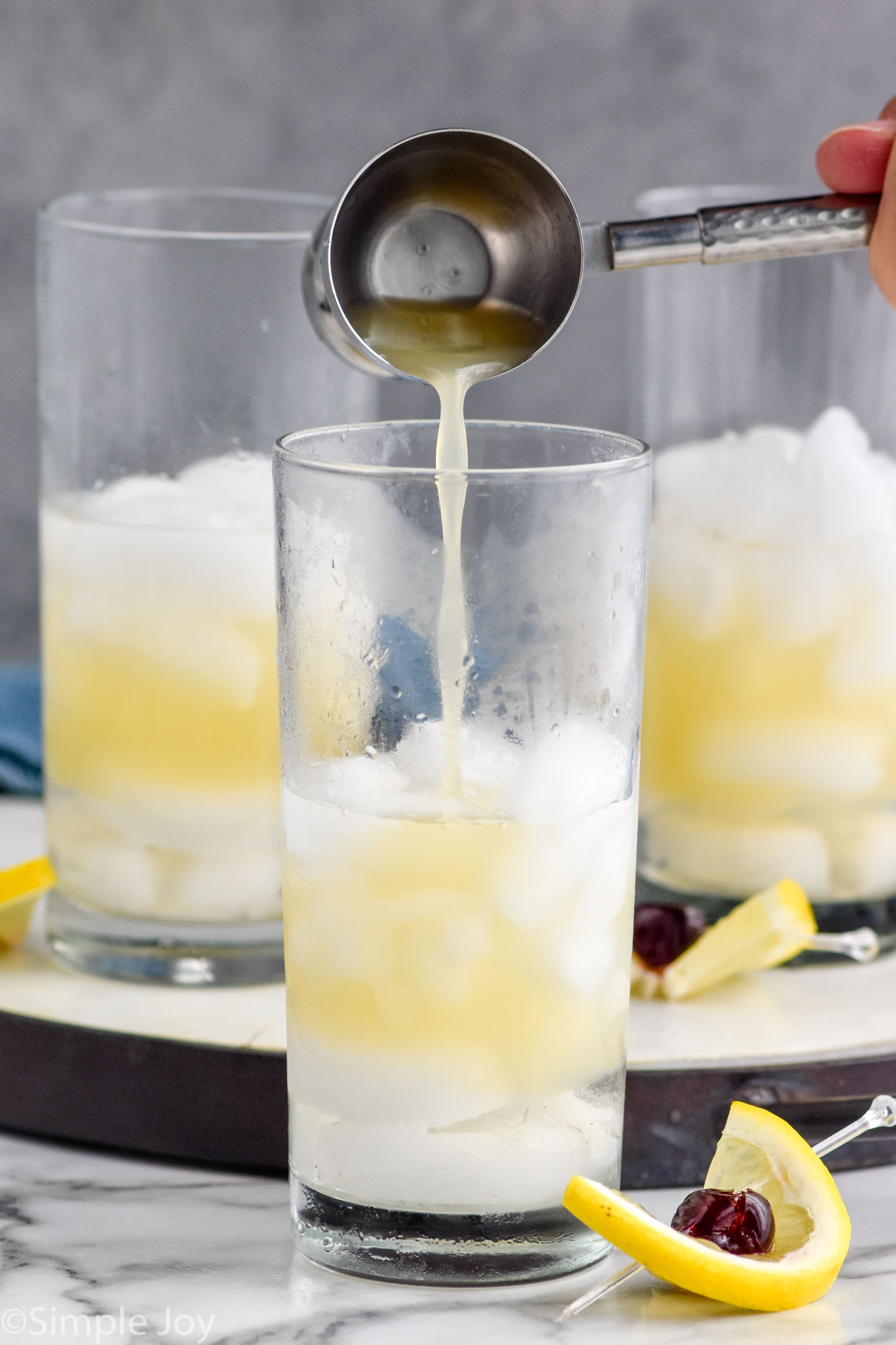 Man's hand pouring cocktail jigger of lemon juice into a glass of ice and Tom Collins ingredients