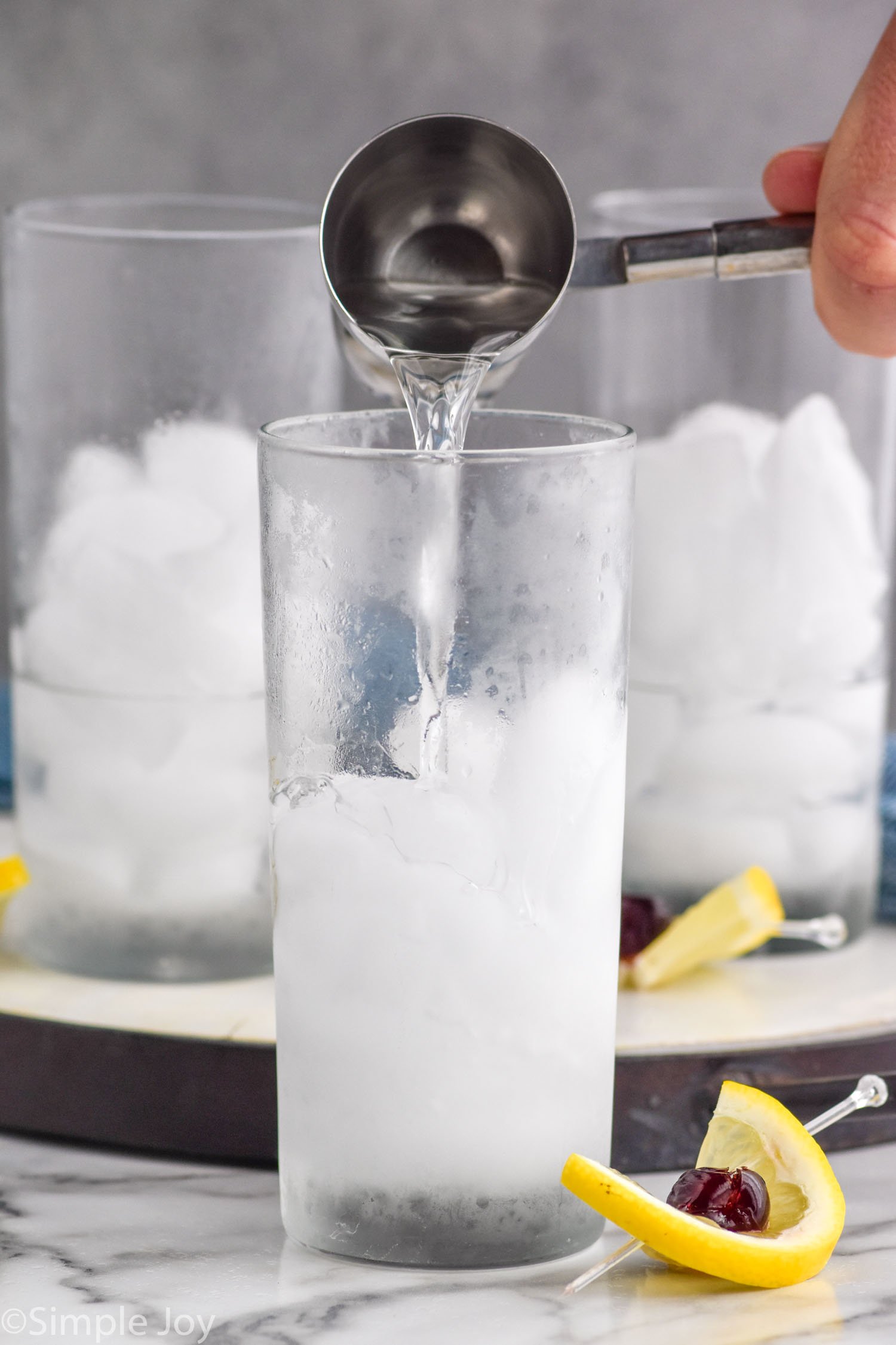 man's hand pouring cocktail jigger of gin into a glass of ice to make a tom collins