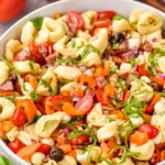Side view of a bowl of Tortellini Pasta Salad. Serving spoons and tomatoes beside bowl. - 9