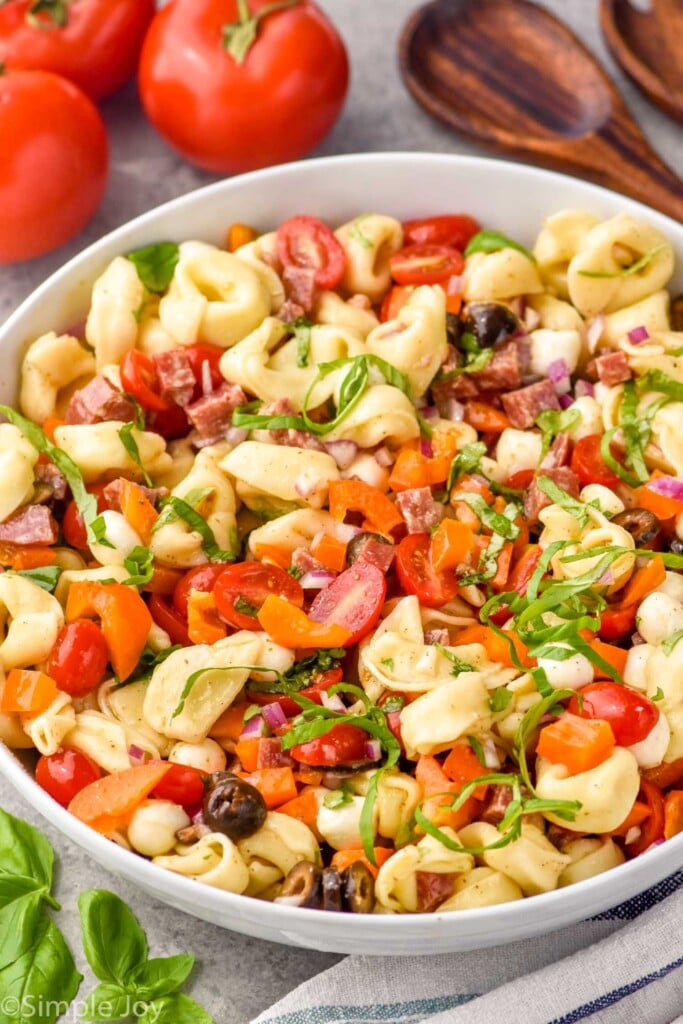 Side view of a bowl of Tortellini Pasta Salad. Serving spoons and tomatoes beside bowl.