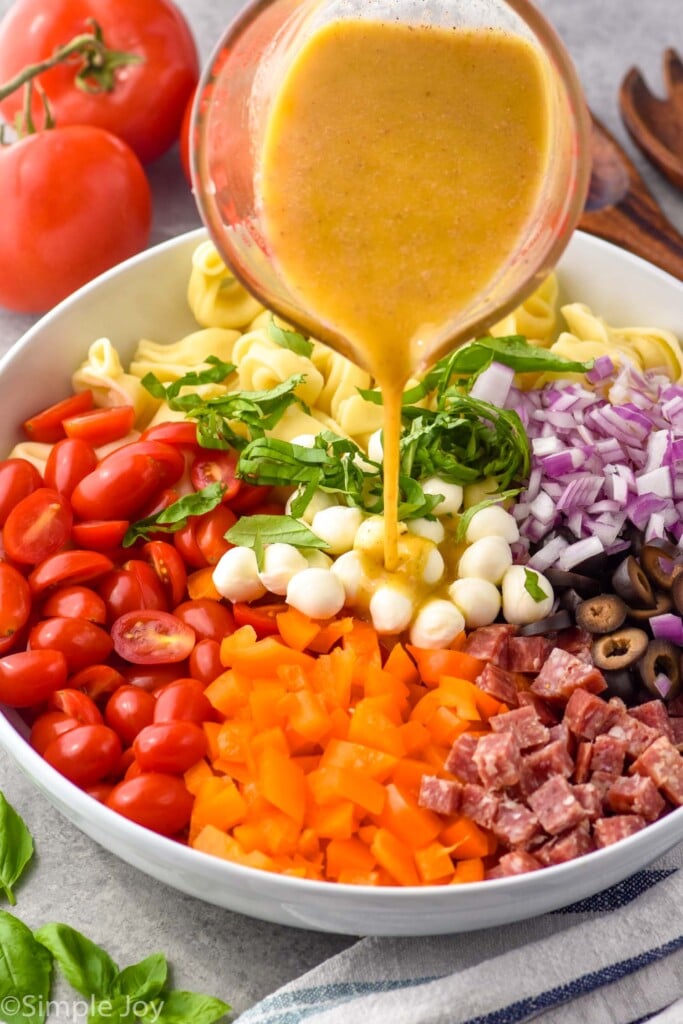 Photo of glass measuring cup of dressing being poured over bowl of ingredients for Tortellini Pasta Salad recipe.