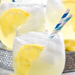 Side view of Vodka Lemonade garnished with lemon slice and straws for drinking. More glasses of Vodka Lemonade in the background.