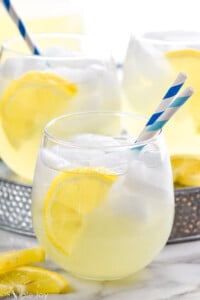 Side view of Vodka Lemonade garnished with lemon slice and straws for drinking. More glasses of Vodka Lemonade in the background.