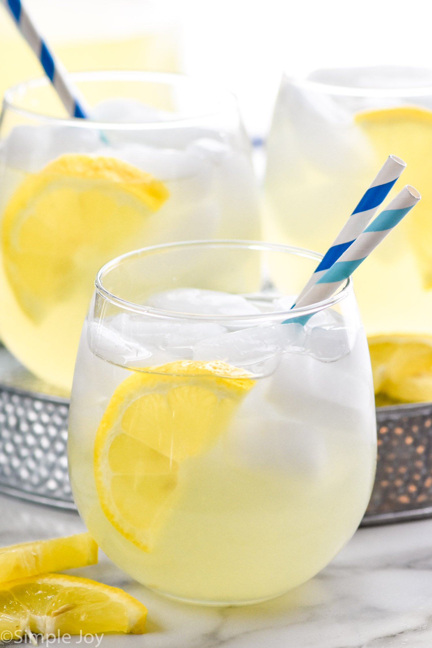 Side view of Vodka Lemonade garnished with lemon slice and straws for drinking. More glasses of Vodka Lemonade in the background.