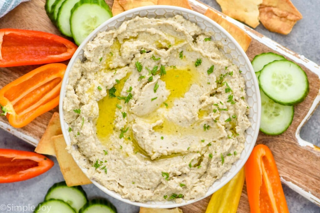 Overhead view of a bowl of Baba Ganoush on a serving board surrounded by fresh cucumbers, peppers, and cracks for serving.