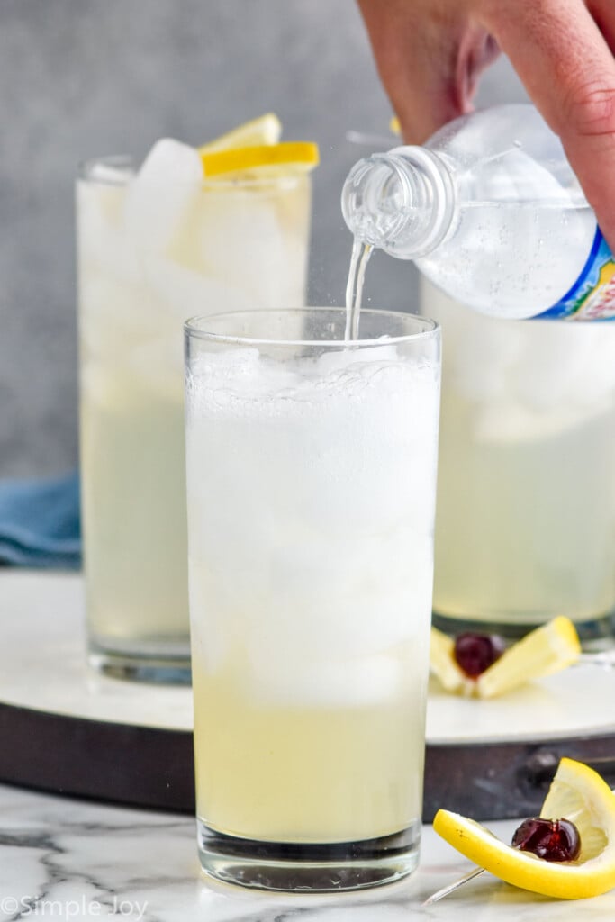 man's hand pouring bottle of soda water into a glass of tom collins ingredients. Lemon and cherry sitting beside