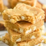 Side view of a stack of Blondie brownies with a bite taken out of the top brownie. White chocolate chips beside stack.