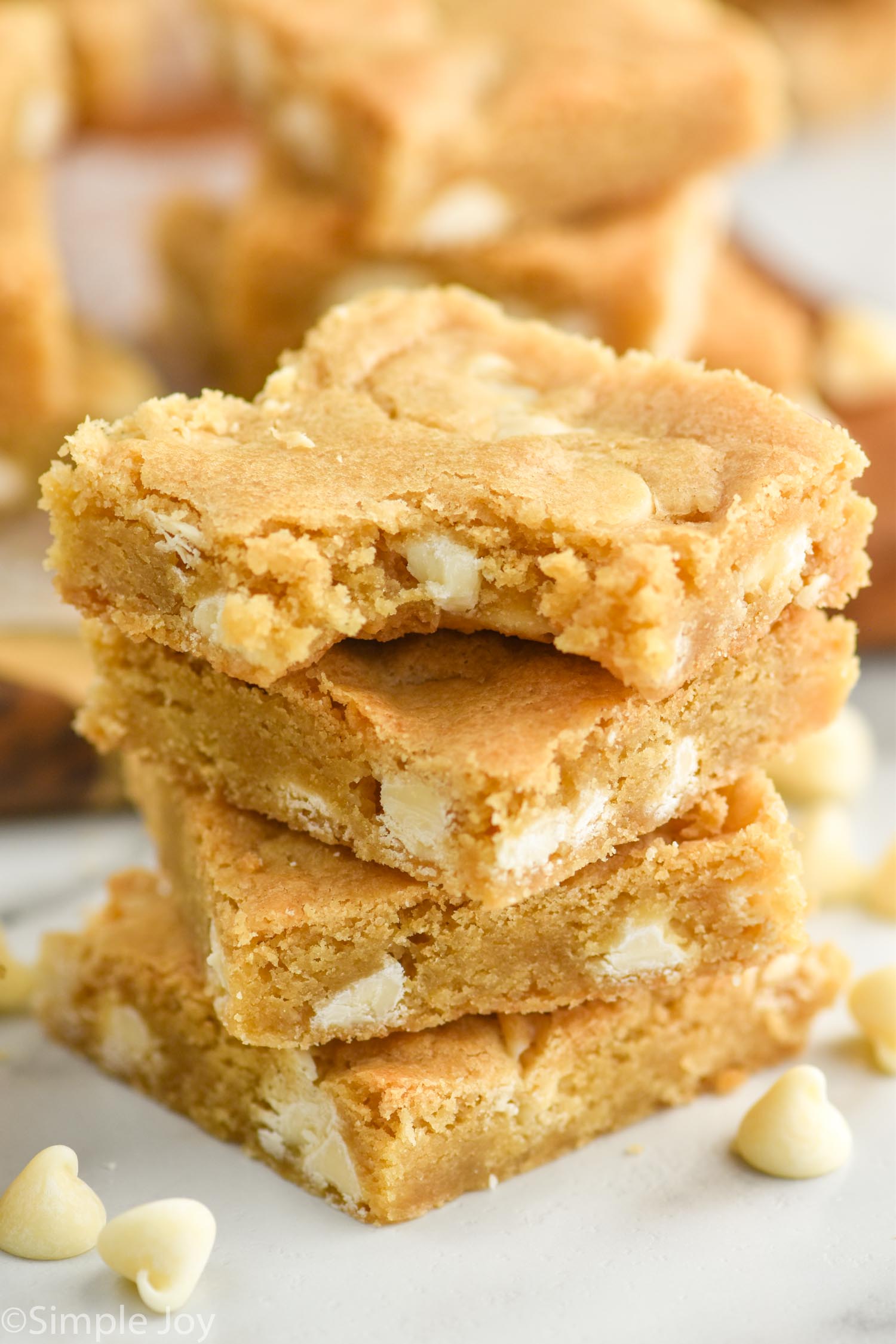 Side view of a stack of Blondie brownies with a bite taken out of the top brownie. White chocolate chips beside stack.