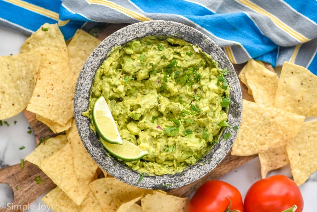 Overhead photo of a bowl of Easy Guacamole Recipe garnished with lime wedges. Chips and tomatoes next to bowl.