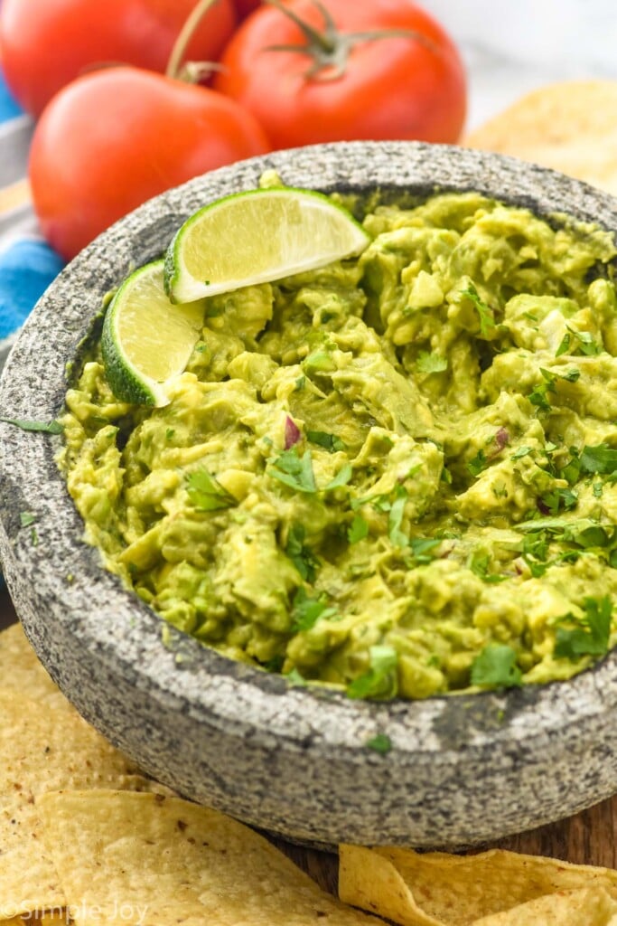 Photo of a bowl of Easy Guacamole Recipe garnished with lime wedges. Chips and tomatoes on the side.