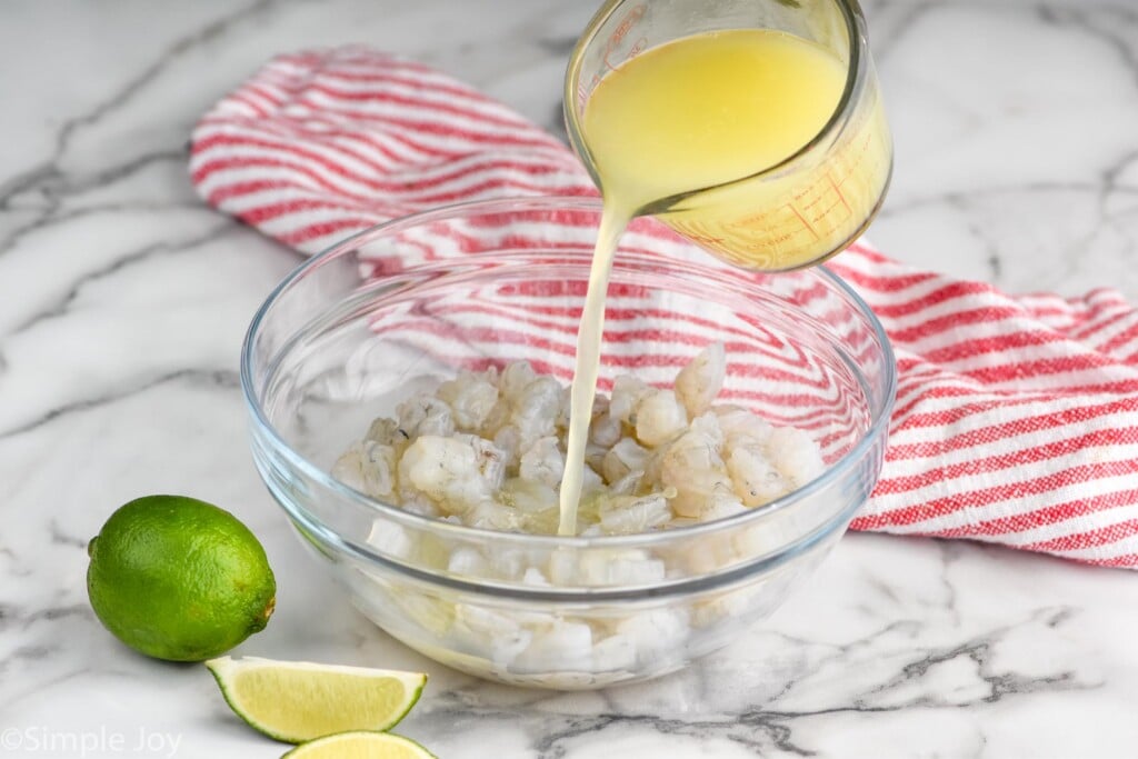 glass measuring cup pouring lemon and lime juices into a bowl of shrimp to make shrimp ceviche. Lime and lime slices sit beside.