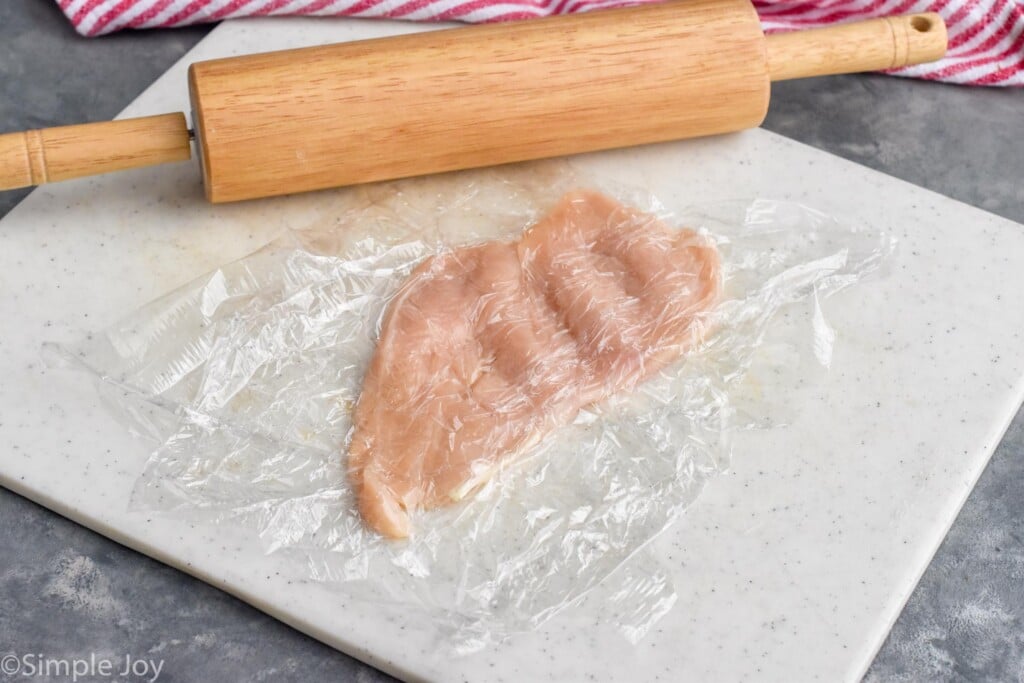 chicken cutlet in plastic wrap on cutting board to make Chicken Cordon Bleu with rolling pin in background