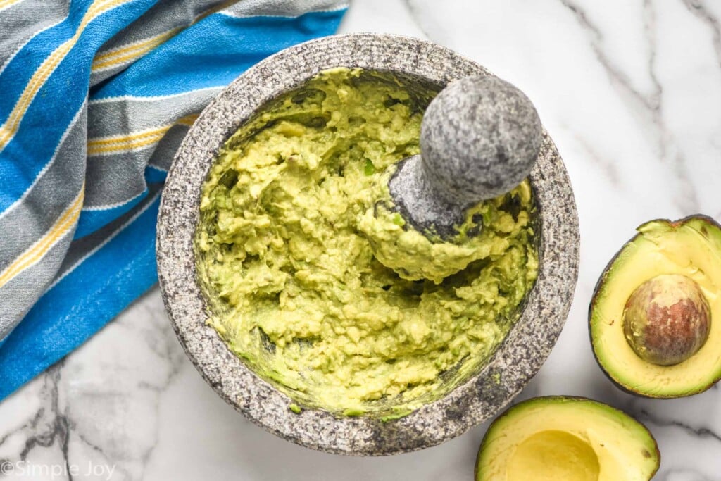 Overhead photo of a mortar and pestle of Easy Guacamole Recipe. Avocado cut in half beside on counter.