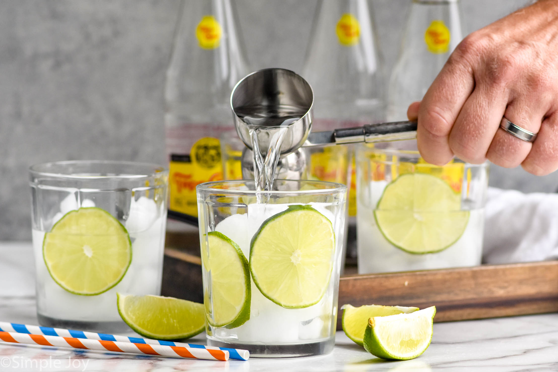 man's hand pouring cocktail jigger of tequila into a glass of ice and slices of lime to make ranch water drink. Ranch water ingredients sitting in background