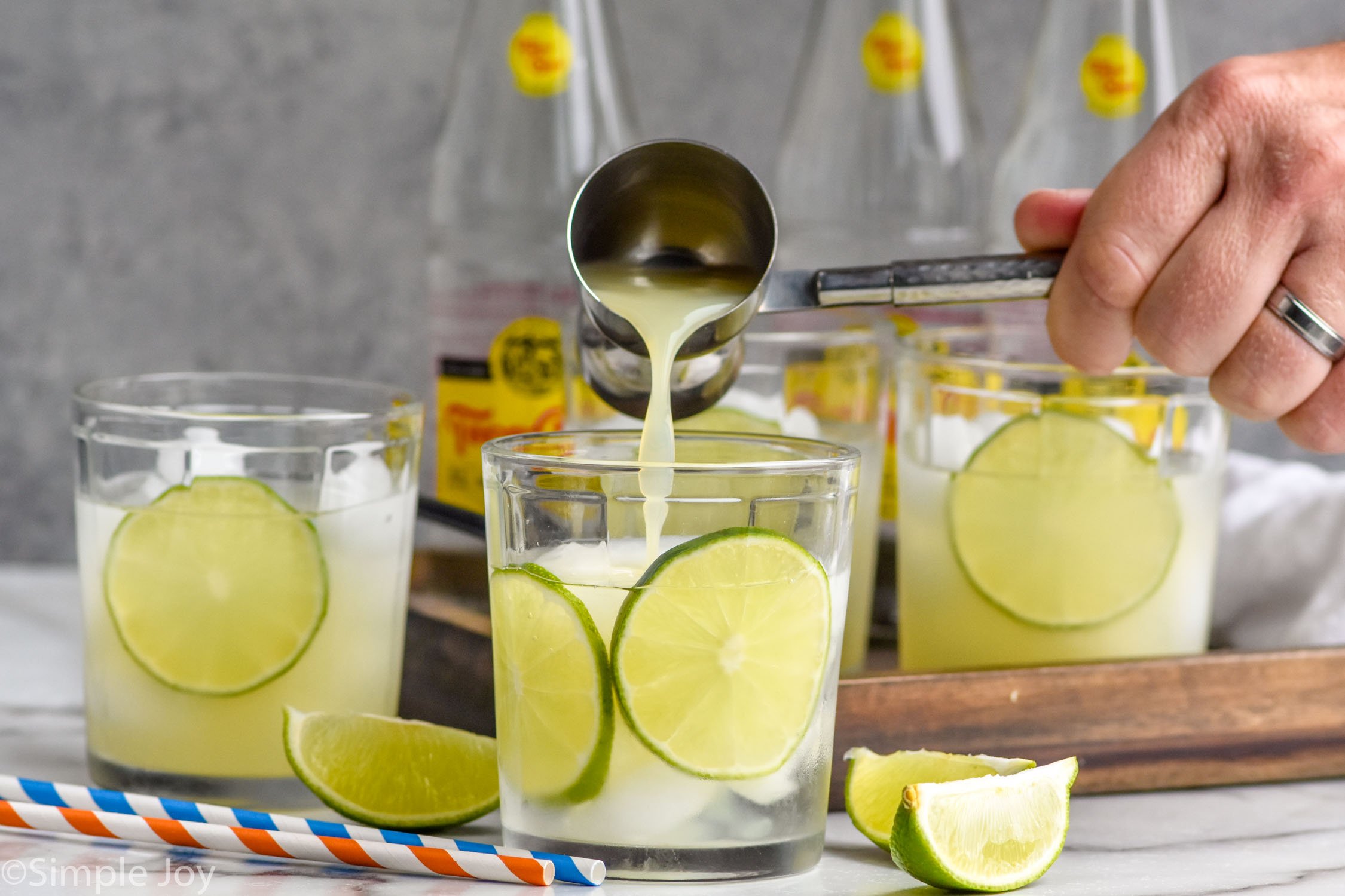 man's hand pouring cocktail jigger of lime juice into a glass of ranch water ingredients and ice.