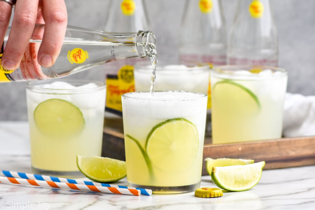 man's hand pouring bottle of topo chico into a glass of ranch water ingredients and ice. Glasses of ranch water and ranch water ingredients sitting in background