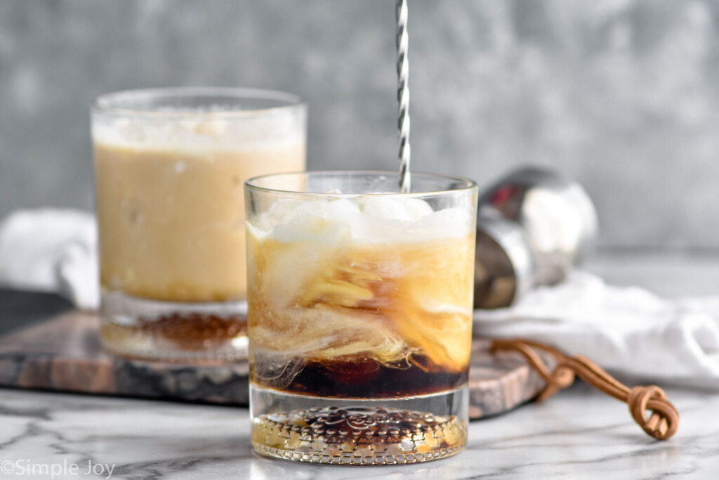 stir stick stirring glass of white russian ingredients. Glass of white russian cocktail sitting in background