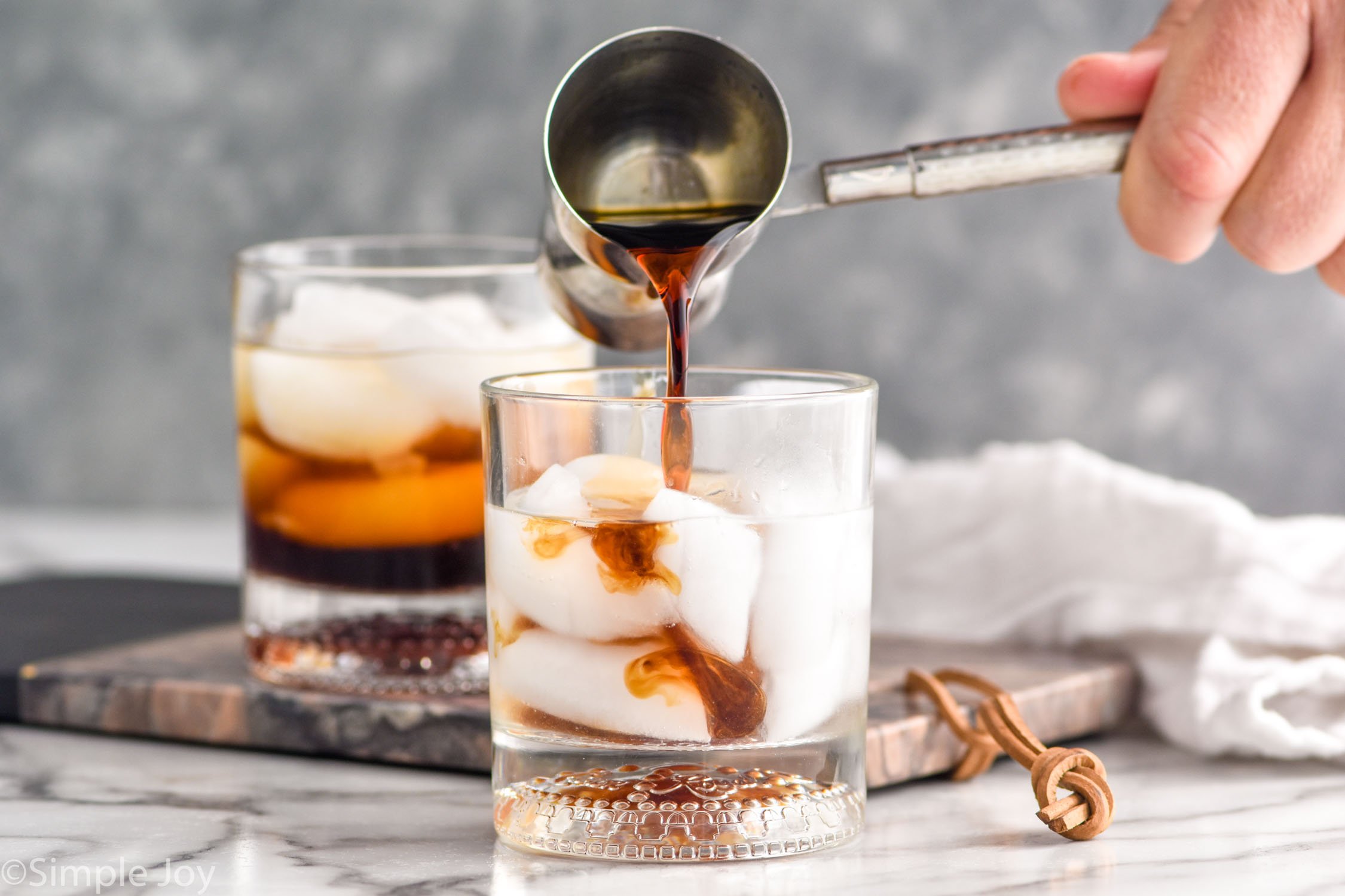 man's hand pouring cocktail jigger of Kahlua into a glass of white russian ingredients. Glass of white russian ingredients sitting in background