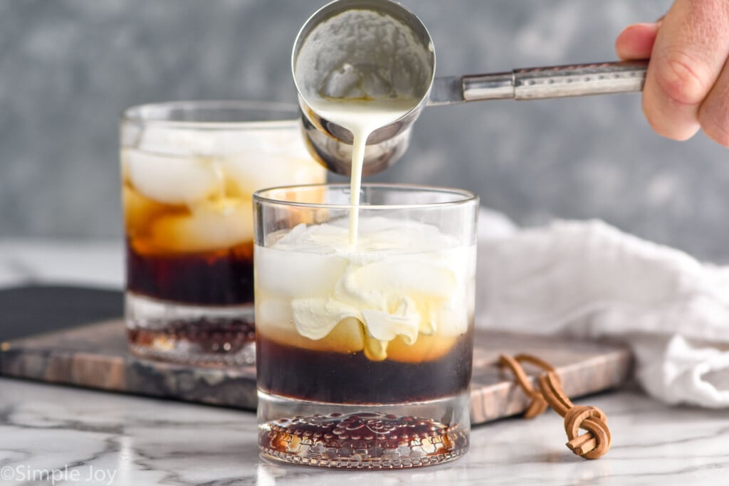 man's hand pouring cocktail jigger of cream into a glass of white russian ingredients and ice. Glass of white russian sitting in background