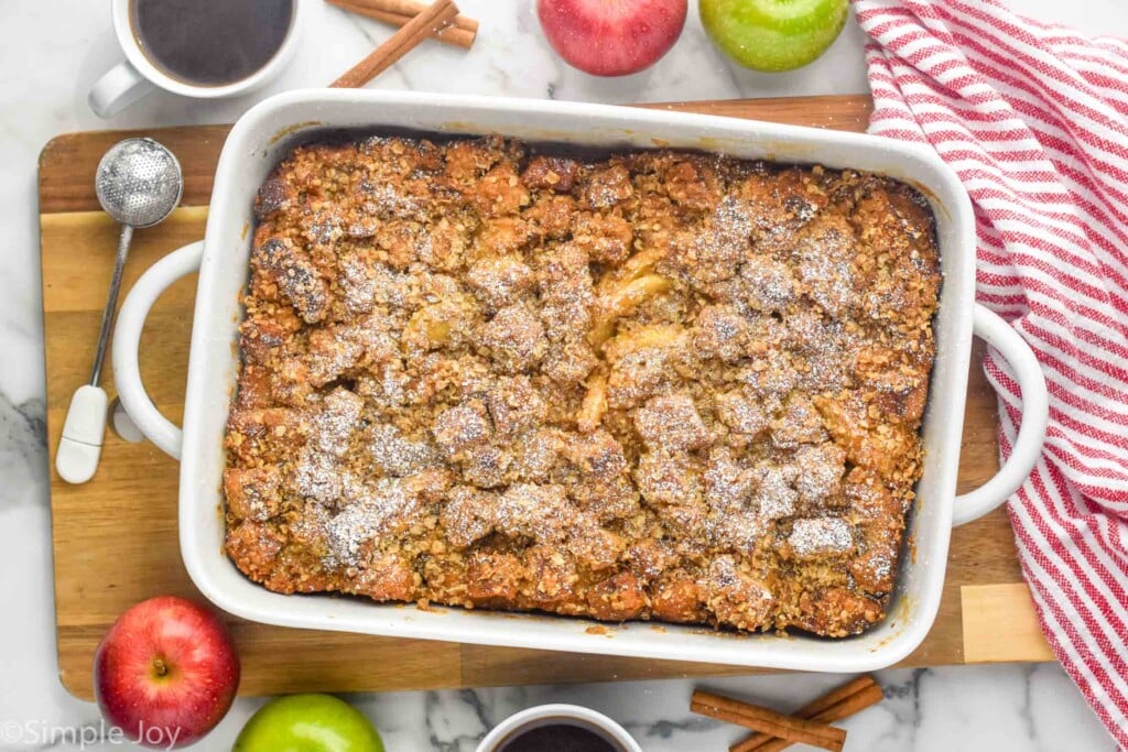 Overhead photo of a baking dish of Apple French Toast Casserole. Apples, cinnamon sticks, cups of coffee, and kitchen utensils beside baking dish,