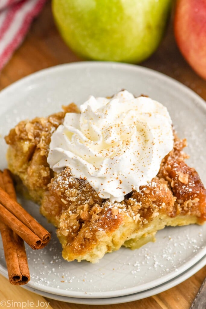 Photo of a piece of Apple French Toast Casserole served on a plate, garnished with whipped cream and cinnamon sticks. Apples beside plate.