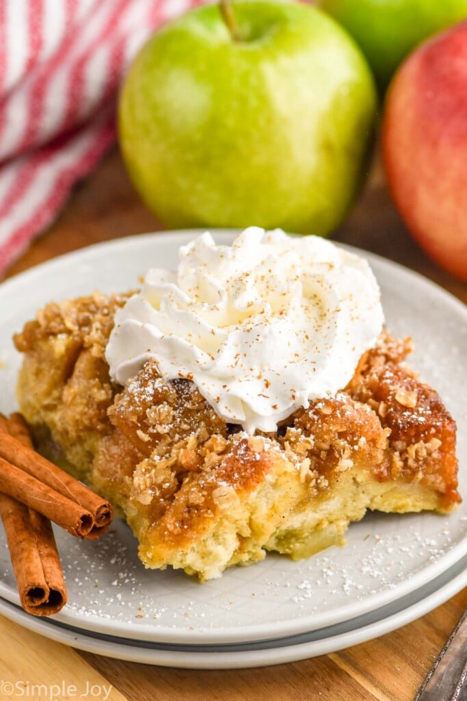 Photo of a piece of Apple French Toast Casserole served on a plate, garnished with whipped cream and cinnamon sticks. Apples beside plate.
