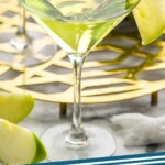 pinterest graphic for appletini. Image shows two martini glasses of Appletini garnished with a green apple slice. Green apple and slices surrounding. Text says "the best appletini simplejoy.com"