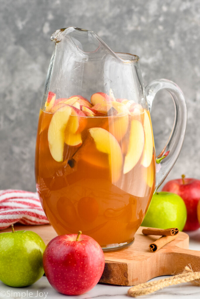 Side view of pitcher of Apple Cider Sangria with apples and cinnamon sticks beside it.
