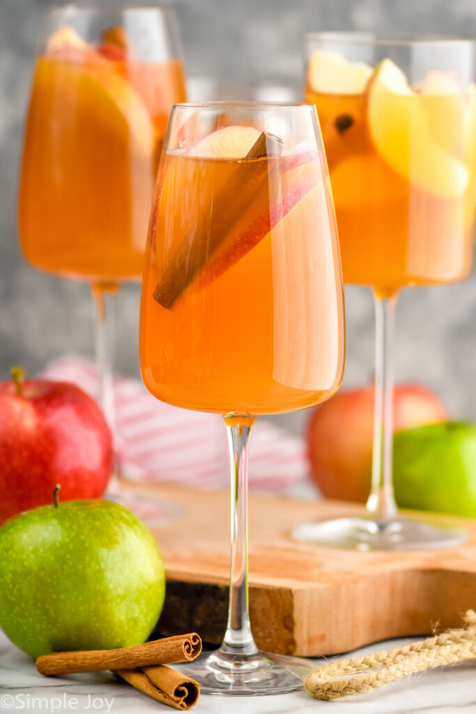 Side view of wine glasses of Apple Cider Sangria garnished with apple slices and cinnamon sticks. Apples and cinnamon sticks beside glasses.
