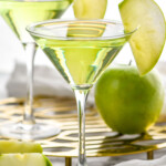 two martini glasses of Appletini garnished with a green apple slice. Green apple and slices surrounding. - 7
