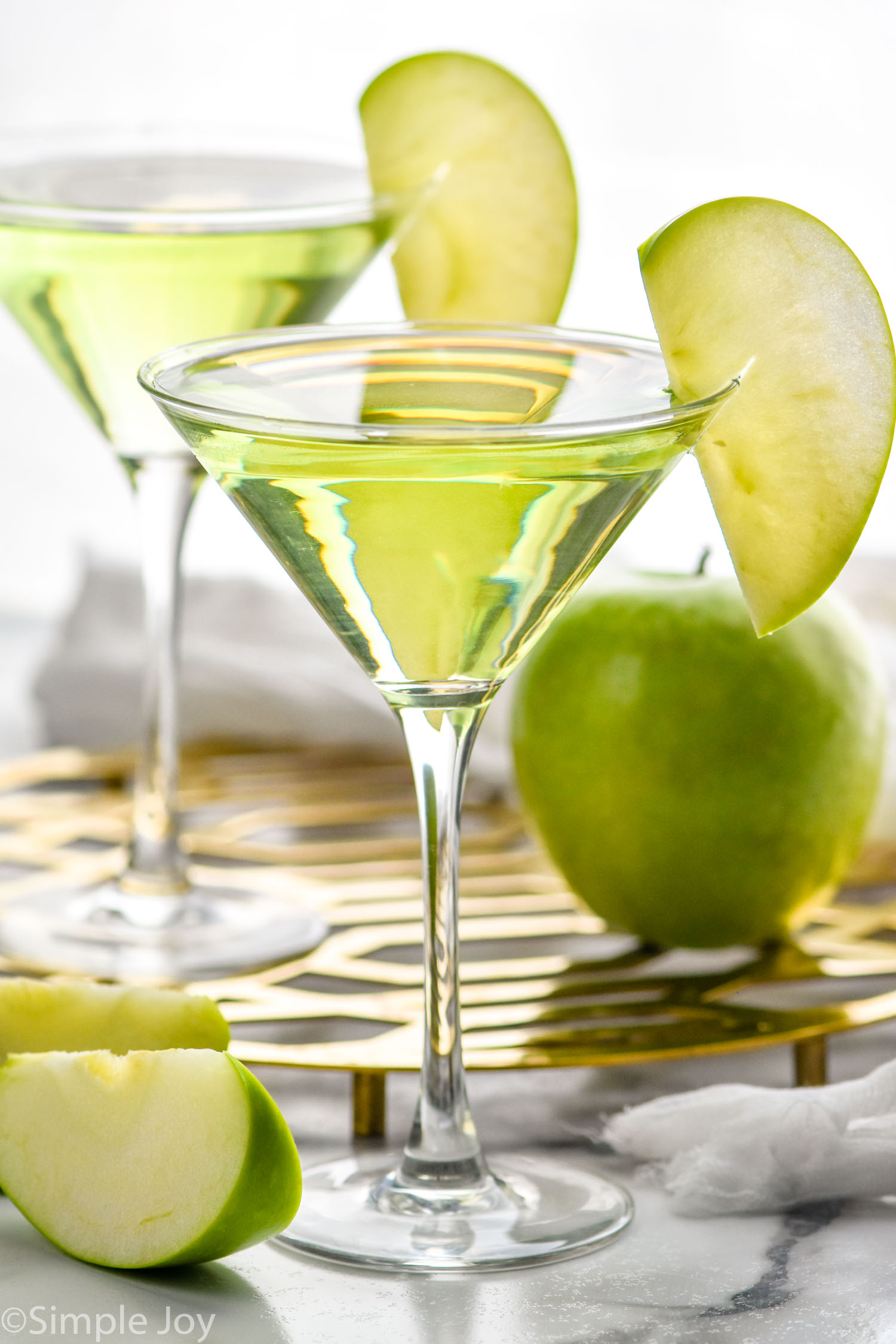 two martini glasses of Appletini garnished with a green apple slice. Green apple and slices surrounding.