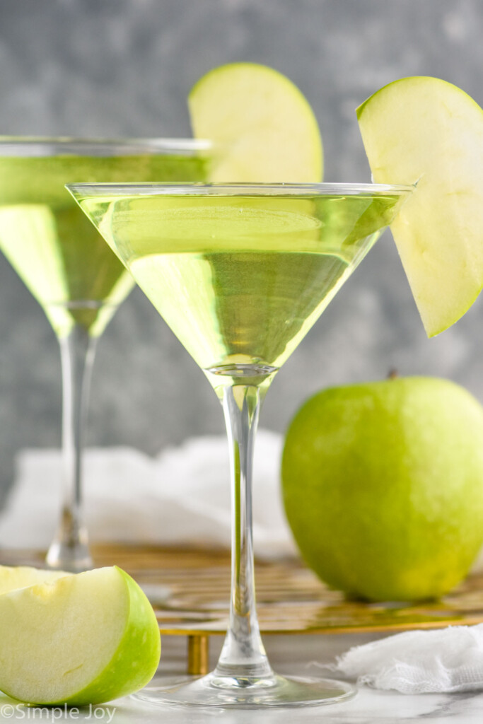 two martini glasses of Appletini garnished with a green apple slice. Green apple and slices surrounding.