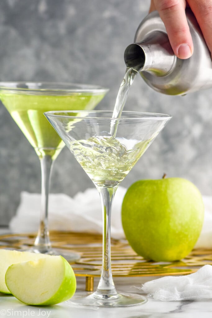 man's hand pouring cocktail shaker of Appletini ingredients into a martini glass. Green apple and glass of Appletini sitting in background