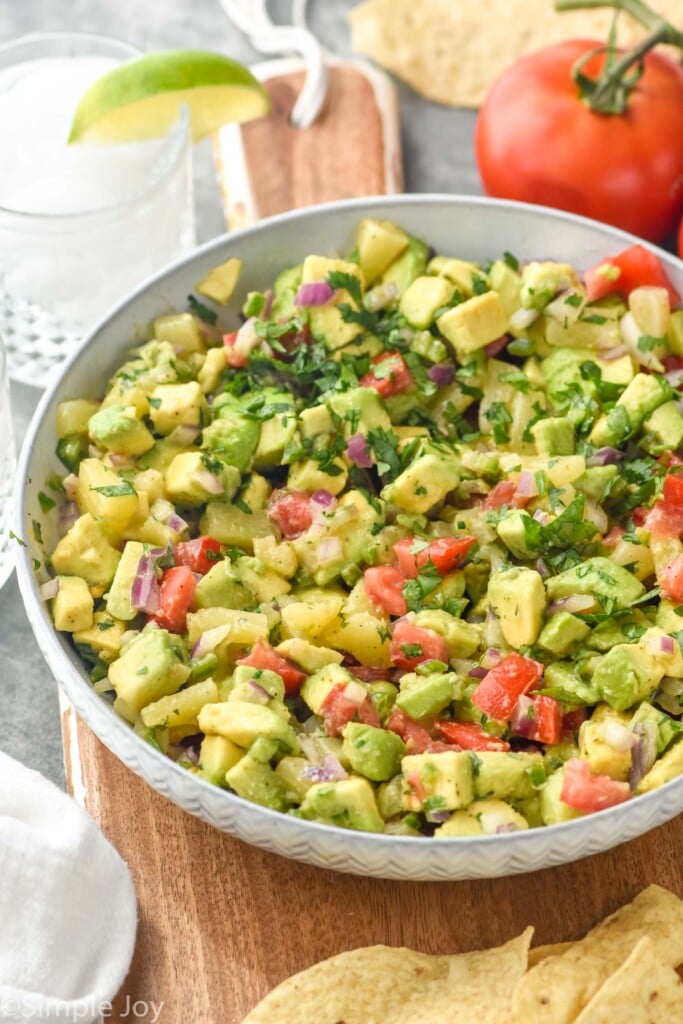 Photo of a bowl of Avocado Salsa. Chips, tomato, and margarita beside bowl.