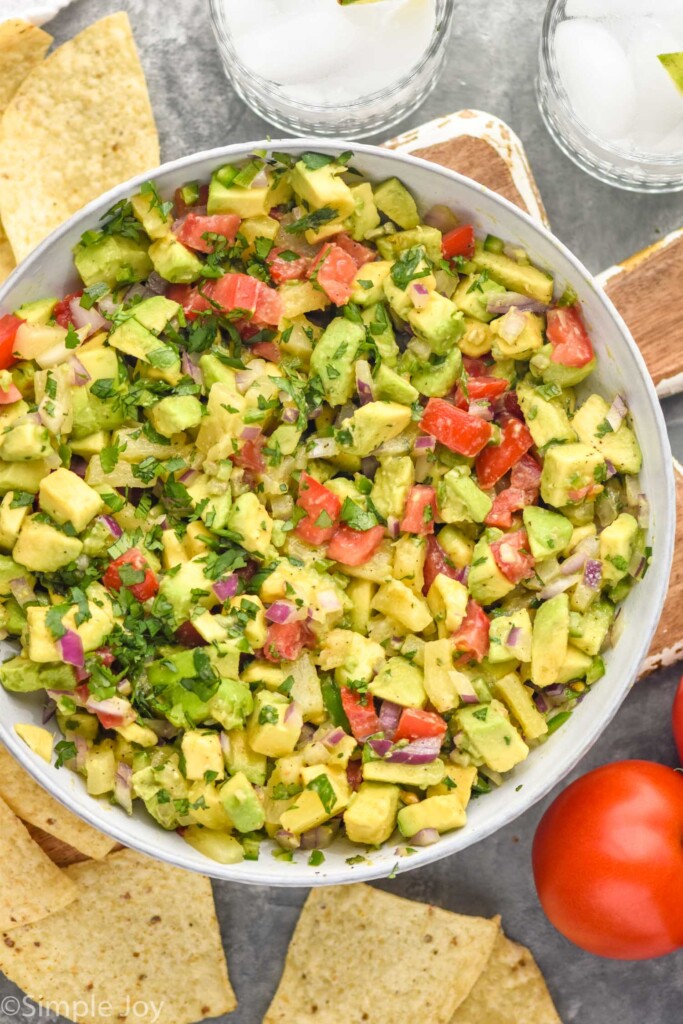 Overhead photo of a bowl of Avocado Salsa with chips, tomatoes, and two glasses beside it