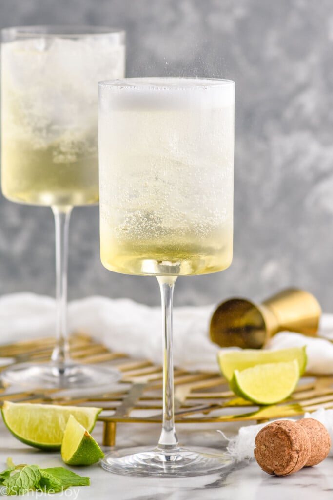 Side view of two wine glasses of Hugo Spritz. Cork, cocktail jigger, and extra lime wedges beside glasses